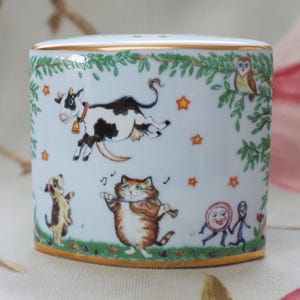 May include: White ceramic cylinder with a gold rim, featuring a whimsical illustration of a cow, cat, dog, owl, and other characters. The design includes stars and green foliage, creating a playful, storybook theme.