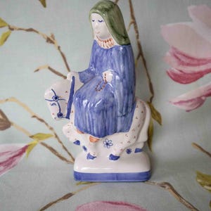 Op de afbeelding: Een keramiek beeldje van een vrouw in een blauwe jurk en een groene hoofdbedekking die op een witte ezel rijdt. De ezel is versierd met blauwe en rode bloemen. Het beeldje staat op een witte basis met een blauwe rand.