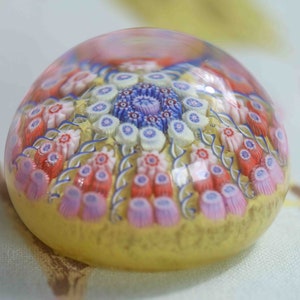 May include: A glass paperweight with a yellow base and a multi-colored floral design. The design features pink, red, blue, and white flowers.