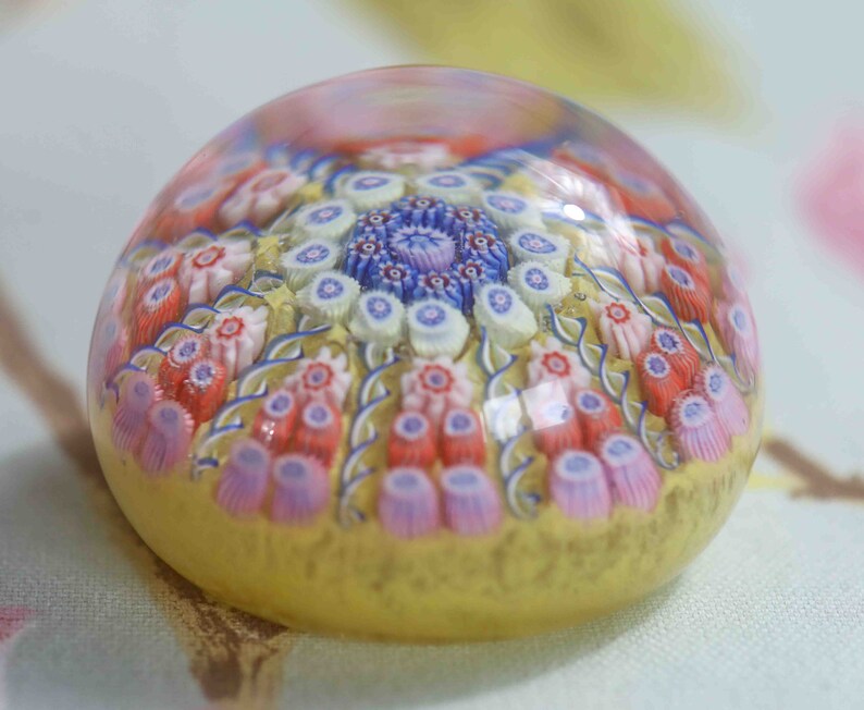 May include: A glass paperweight with a yellow base and a multi-colored floral design. The design features pink, red, blue, and white flowers.