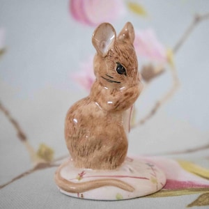May include: A brown ceramic figurine of a mouse holding a small book. The mouse is sitting on a white base with pink and green floral details.