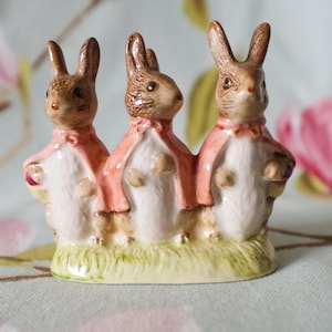 May include: A ceramic figurine featuring three brown and white rabbits standing side by side on a green base. Each rabbit wears a pink cape tied at the neck with a small bow. The rabbits have detailed facial features and are positioned in a row.