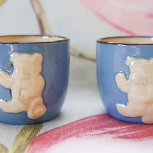 Peut inclure: Deux tasses en céramique bleues avec un motif d'ours en peluche beige clair. Les tasses ont une forme arrondie et un bord marron. L'ours en peluche est en relief.