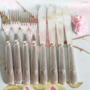 Può includere: Set di otto posate argentate, tra cui coltelli e forchette, con un motivo a diamante decorativo sui manici. Le posate sono disposte su una tovaglia floreale blu chiaro e rosa.