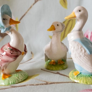 May include: Three ceramic duck figurines standing on green bases. The left duck wears a blue bonnet and pink shawl. The center duck is plain white with an orange beak. The right duck wears a blue jacket. All have orange feet and beaks.