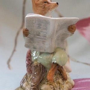May include: A ceramic figurine of a fox reading a newspaper. The fox is depicted with brown fur and is wearing a green outfit. The newspaper is white with gray text. The figurine is sitting on a brown base.