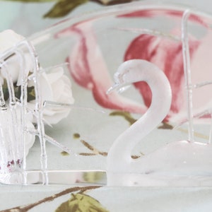 May include: A clear glass swan figurine with a crystal design. The swan is sitting on a clear glass base with a small label that reads "Kosta Boda Sweden Full Lead Crystal".