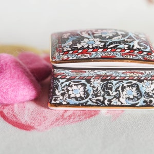 Peut inclure: Une petite boîte carrée décorative avec un motif floral noir, blanc et bleu. La boîte a une bordure dorée et est posée sur un tissu floral rose et blanc.