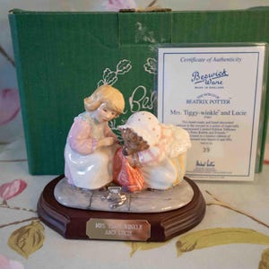 May include: A porcelain figurine of two girls from Beatrix Potter's "Mrs. Tiggy-Winkle" story. The figurine depicts Mrs. Tiggy-Winkle, a hedgehog, and Lucie, a young girl, in a charming scene. The figurine is on a wooden base with a gold plaque that reads "Mrs. Tiggy-Winkle and Lucie".