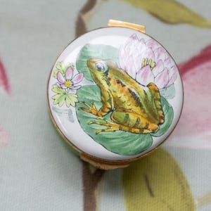 May include: A round, white trinket box with a gold rim. The lid features a detailed illustration of a frog on a lily pad with a pink lotus flower and small pink flowers. The frog is yellow and green.