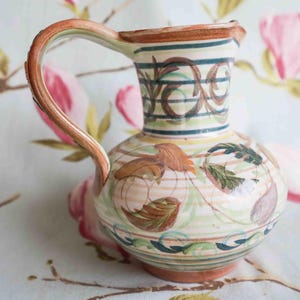Può includere: Brocca in ceramica dipinta a mano con corpo arrotondato e manico curvo. Presenta una base color crema con disegni floreali e di foglie marroni, verdi e blu. Strisce orizzontali e volute decorative adornano il collo.