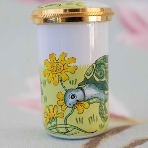 May include: A small, cylindrical container with a gold-colored rim and lid. The container is white with a yellow and green painted scene of a turtle and yellow flowers. The lid is green and white.