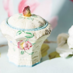 May include: A white ceramic sugar bowl with a floral design. The bowl has a lid with a floral knob and a blue trim around the top.