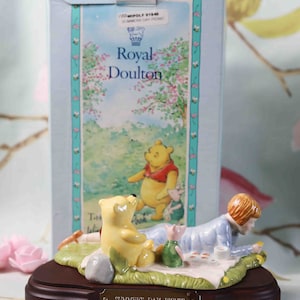 May include: A porcelain figurine of Winnie the Pooh and Piglet having a picnic with a wooden base. The base has a gold plaque that reads "Summer's Day Picnic".