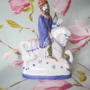 Op de afbeelding: Een keramiek beeldje van een man die op een paard rijdt. De man draagt een blauwe robe en een rode tulband. Het paard is wit met blauwe en witte details. Het beeldje is versierd met blauwe en witte bloemmotieven.