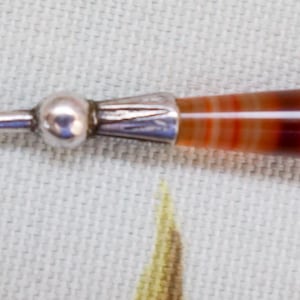 Può includere: Un uncinetto vintage con un gancio in metallo argentato e un manico in agata a bande nei toni del rosso, marrone e bianco. L'uncinetto presenta un piccolo dettaglio rotondo in metallo argentato dove il manico incontra il gancio.