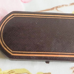 Puede incluir: Una caja rectangular marr&oacute;n con un borde dorado. La caja tiene una superficie texturizada y est&aacute; cerrada.