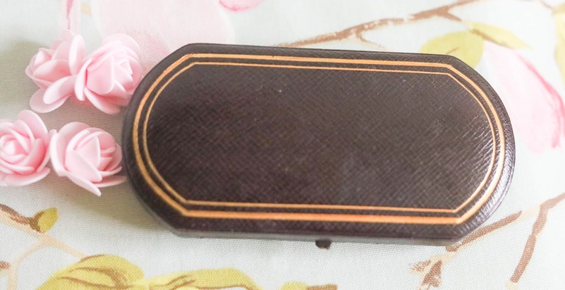 Puede incluir: Una caja rectangular marr&oacute;n con un borde dorado. La caja tiene una superficie texturizada y est&aacute; cerrada.