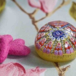 May include: A glass paperweight with a colorful, intricate design of flowers and swirls. The paperweight is sitting on a floral patterned fabric with two pink felt hearts.