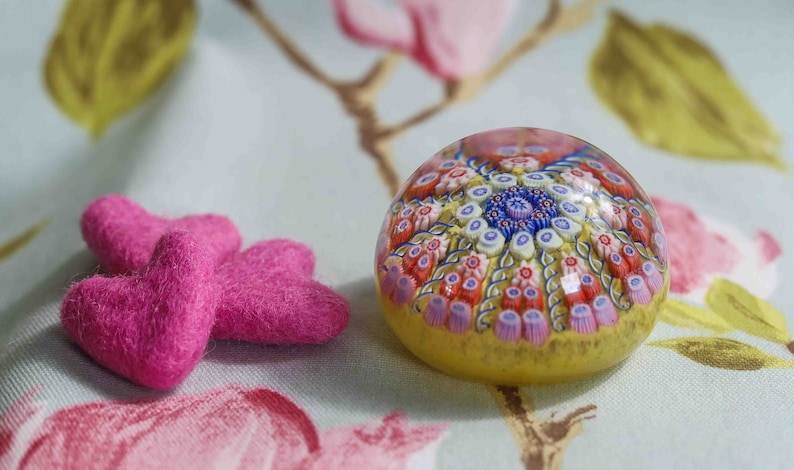 May include: A glass paperweight with a colorful, intricate design of flowers and swirls. The paperweight is sitting on a floral patterned fabric with two pink felt hearts.