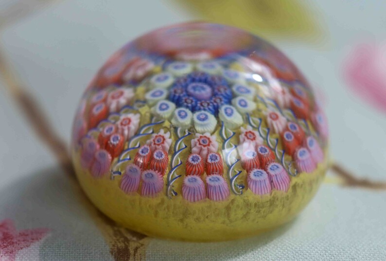 May include: A glass paperweight with a colorful floral design. The paperweight is round and has a clear glass base. The flowers are in shades of pink, blue, and white.