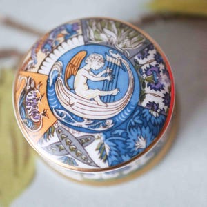 May include: A round porcelain trinket box featuring a colourful floral and cherub design. The cherub is playing a harp and is surrounded by blue, yellow and green floral patterns.