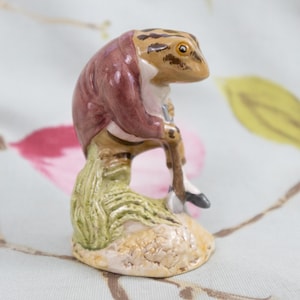 Könnte beinhalten: Eine Keramikfigur einer Froschfigur in einer bordeauxfarbenen Jacke, die einen Spazierstock hält. Der Frosch ist beige und braun mit einem weißen Kragen. Die Figur steht auf einem kleinen, strukturierten Sockel mit grünen und beigen Akzenten.