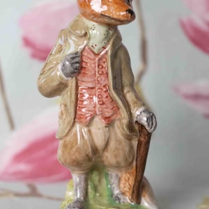 May include: A ceramic figurine of a fox dressed in a tan coat, vest, and breeches, holding a cane. The fox has a brown and black head and is standing on a green base. The figurine is a decorative item.