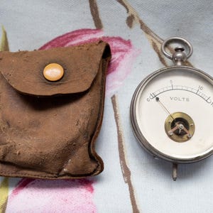 May include: A vintage voltmeter with a white face and a needle indicating voltage, next to a brown leather pouch with a button closure. The voltmeter reads "VOLTS" and has a metal casing.