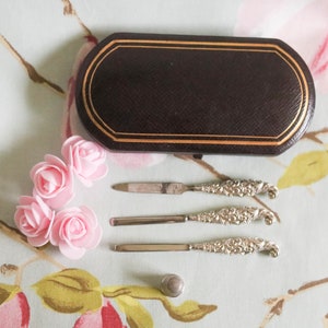 Puede incluir: Un juego de tres herramientas de manicura plateadas con mangos adornados, una peque&ntilde;a caja negra con ribete dorado y cuatro rosas de tela rosa.