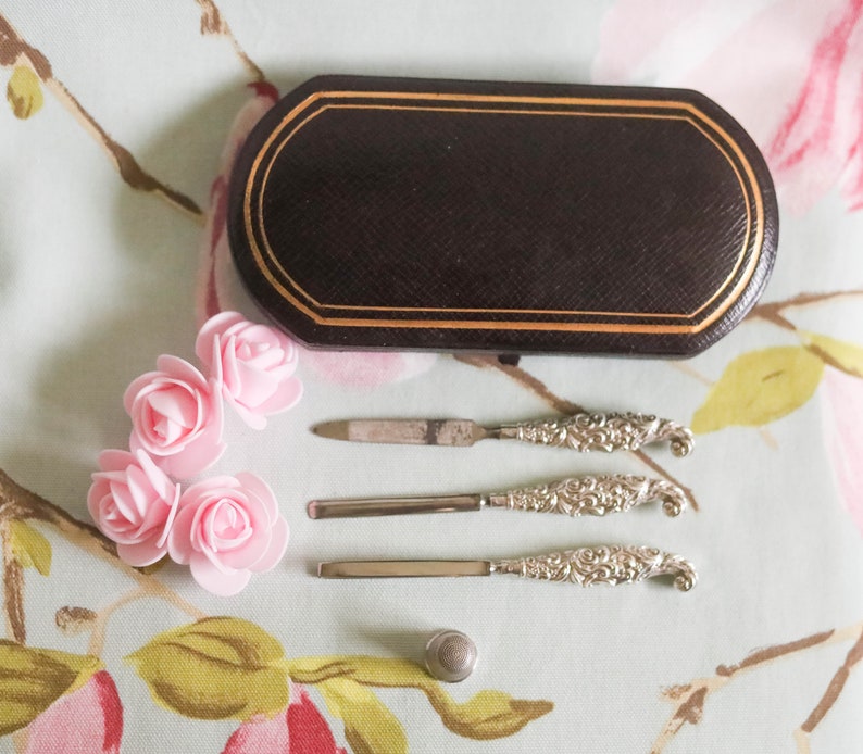 Puede incluir: Un juego de tres herramientas de manicura plateadas con mangos adornados, una peque&ntilde;a caja negra con ribete dorado y cuatro rosas de tela rosa.