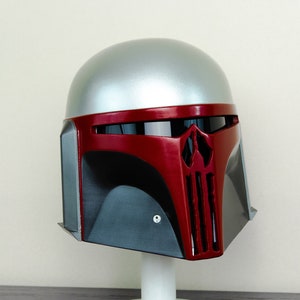 May include: A silver and red Mandalorian helmet with a black visor. The helmet is on a white display stand.