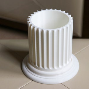 Op de afbeelding: Witte keramische plantenpot in de vorm van een gegroefde kolom. De pot heeft een gladde, afgeronde bovenkant en een basis met een decoratieve rand.