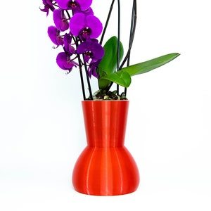 Puede incluir: Una planta de orquídea púrpura vibrante en un jarrón de cerámica naranja brillante. El jarrón tiene una base redonda y un cuello estrecho.