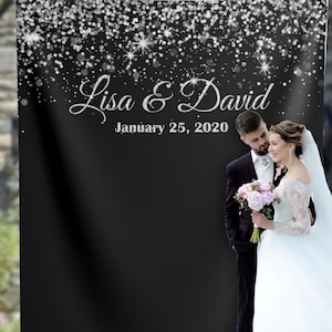 May include: A black backdrop with silver glitter falling from the top. The text "Lisa & David" and "January 25, 2020" is written in silver glitter. A bride and groom are standing in front of the backdrop. The bride is wearing a white wedding dress and the groom is wearing a black suit.
