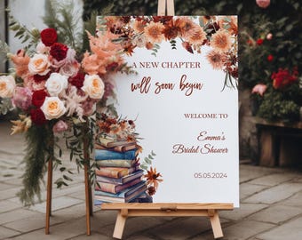 May include: A white sign with a floral design and the text "A new chapter will soon begin. Welcome to Emma's Bridal Shower 05.05.2024". The sign is propped up on an easel.