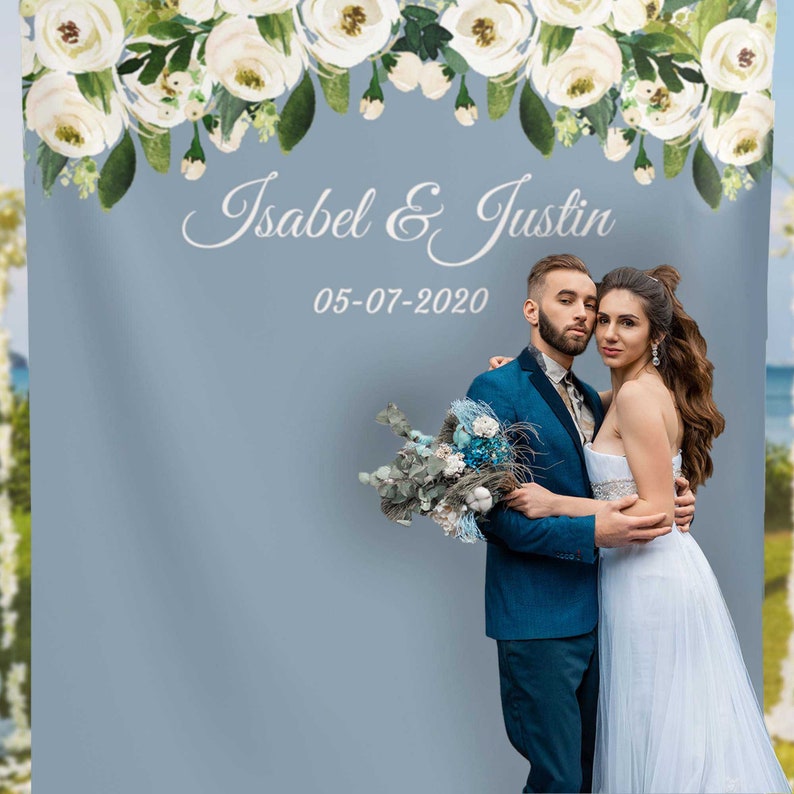 May include: A wedding backdrop with a floral garland at the top, a light blue background, and the text "Isabel & Justin 05-07-2020". A couple is standing in front of the backdrop, the woman in a white dress and the man in a blue suit.