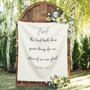 May include: A wooden wedding arch with a cream-colored banner featuring the initials "J&A" and a Bible verse. The arch is decorated with white and green flowers and greenery. The text on the banner reads, "The Lord hath done great things for us; whereof we are glad." Psalm 126:3.
