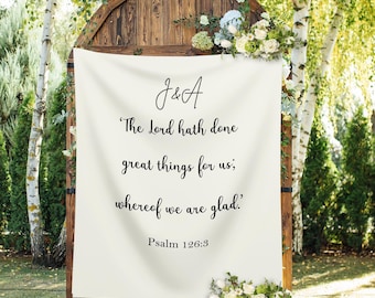 May include: A wooden wedding arch with a cream-colored banner featuring the initials "J&A" and a Bible verse. The arch is decorated with white and green flowers and greenery. The text on the banner reads, "The Lord hath done great things for us; whereof we are glad." Psalm 126:3.