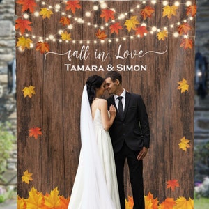 May include: A wedding backdrop with a rustic wood background, autumn leaves, and fairy lights. The text "fall in love" and "TAMARA & SIMON" is printed on the backdrop.