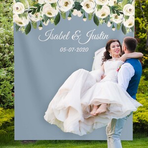May include: A wedding backdrop with a light blue background and a white floral garland at the top. The text "Isabel & Justin" and the date "05-07-2020" are printed in white on the backdrop.