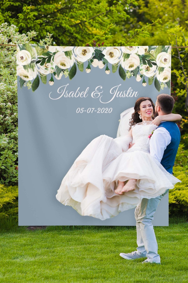 May include: A wedding backdrop with a light blue background and a white floral garland at the top. The text "Isabel & Justin" and the date "05-07-2020" are printed in white on the backdrop.