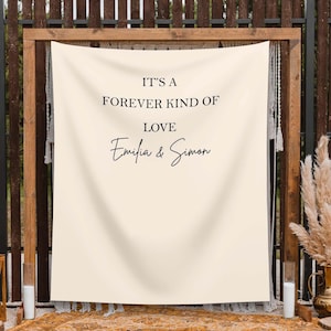 May include: A cream-coloured wedding backdrop featuring the text "IT'S A FOREVER KIND OF LOVE Emilia & Simon" in black script. The backdrop is framed by a wooden structure and decorated with macrame and pampas grass, creating a romantic setting.