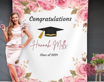 May include: A white graduation backdrop with pink floral designs. The text "Congratulations" is at the top, followed by a black graduation cap with a gold tassel. The name "Hannah Mills" is written in cursive script, and the text "Class of 2024" is below.