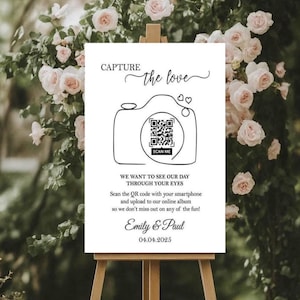 May include: A white wedding sign on an easel with the text "CAPTURE the love." The sign features a camera graphic with a QR code and the words "SCAN ME." It also includes the text "WE WANT TO SEE OUR DAY THROUGH YOUR EYES" and the names "Emily & Paul" with the date 04.04.2025.