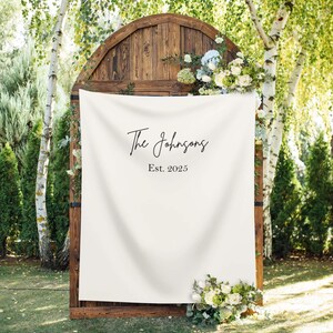 May include: A wooden wedding arch with a cream fabric banner. The banner displays the text "The Johnsons Est. 2025" in black script. The arch is decorated with white and blue flowers and greenery.