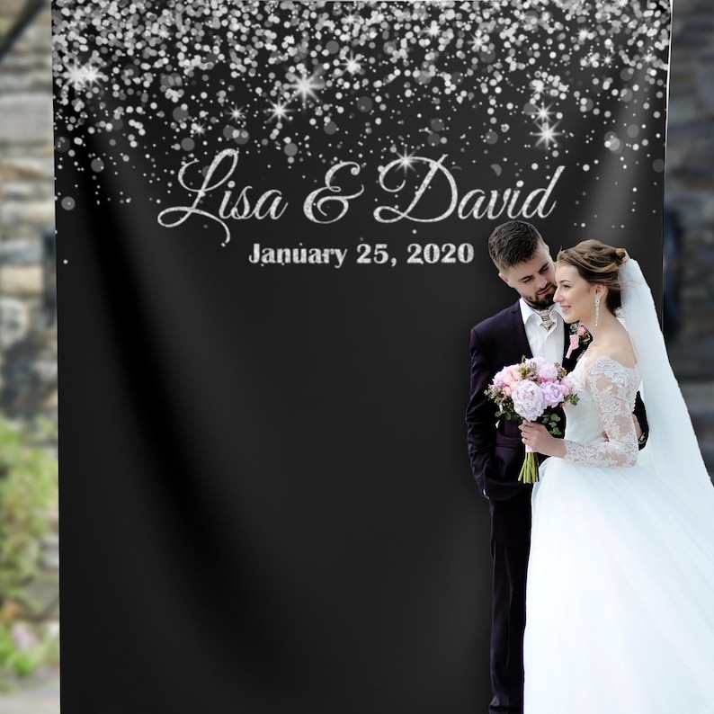 May include: A black backdrop with silver glitter falling from the top. The text "Lisa & David" and "January 25, 2020" is written in silver glitter. A bride and groom are standing in front of the backdrop. The bride is wearing a white wedding dress and the groom is wearing a black suit.