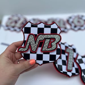 Puede incluir: Un recorte en forma de escudo con un patr&oacute;n de cuadros blancos y negros y un borde rojo. Las letras NB se muestran en una fuente plateada con textura de placa de diamante y un contorno rojo, superpuestas en el centro del dise&ntilde;o del escudo.