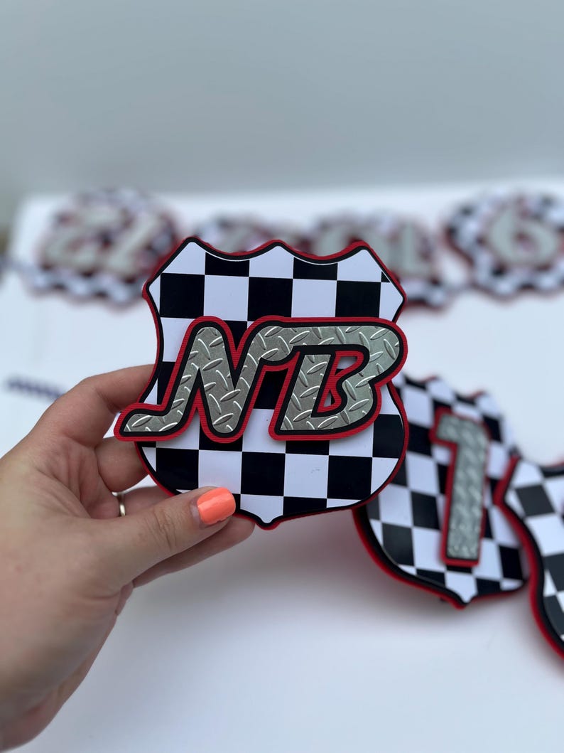 Puede incluir: Un recorte en forma de escudo con un patr&oacute;n de cuadros blancos y negros y un borde rojo. Las letras NB se muestran en una fuente plateada con textura de placa de diamante y un contorno rojo, superpuestas en el centro del dise&ntilde;o del escudo.
