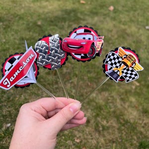 May include: A hand holds several decorative toppers featuring a red race car, a checkered flag, and a metal plate design. The toppers are on clear sticks, with a red and black circular border. One topper has the name "Jameson" on it.